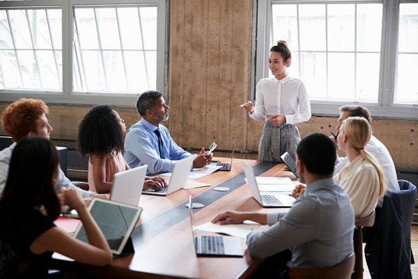 Female-manager-stands-addressing-team-at-board-meeting-IST-904596172